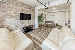 A modern living room with a stone wall, a flat-screen TV, a foosball table, and two beige sofas. - Waterfront house in Virginia Beach with amazing views, private pool & Hot Tub (Virginia Beach)