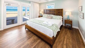 A bedroom with a large bed, a ceiling fan, a nightstand with a lamp, and a view of the beach through a sliding door.