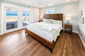 A bedroom with a large bed, a ceiling fan, a nightstand with a lamp, and a view of the beach through a sliding door.