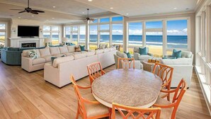 A spacious living room with a large window overlooking the beach, a fireplace, and a dining area with orange chairs.
