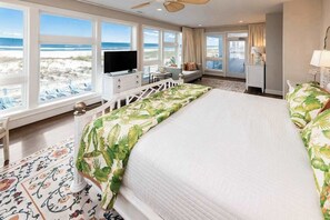 A hotel room with a large bed, a TV, a desk with a chair, a balcony with a view of the beach, and a large window.