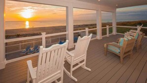 A covered deck with white railings, wooden chairs, and a view of the sunset over the ocean.