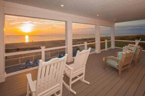 A covered deck with white railings, wooden chairs, and a view of the sunset over the ocean.