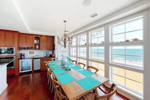 A kitchen with wooden cabinets, a dining table with blue placemats, and a view of the beach through large windows. - Oceanfront corner penthouse with sunrise/sunset views, three pools, & two gyms (Virginia Beach)