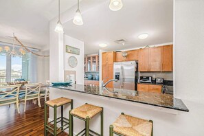A modern kitchen with wooden cabinets, a stainless steel refrigerator, and a bar area with stools. - Condo near the beach & pool with 2 balconies & central AC (Virginia Beach)