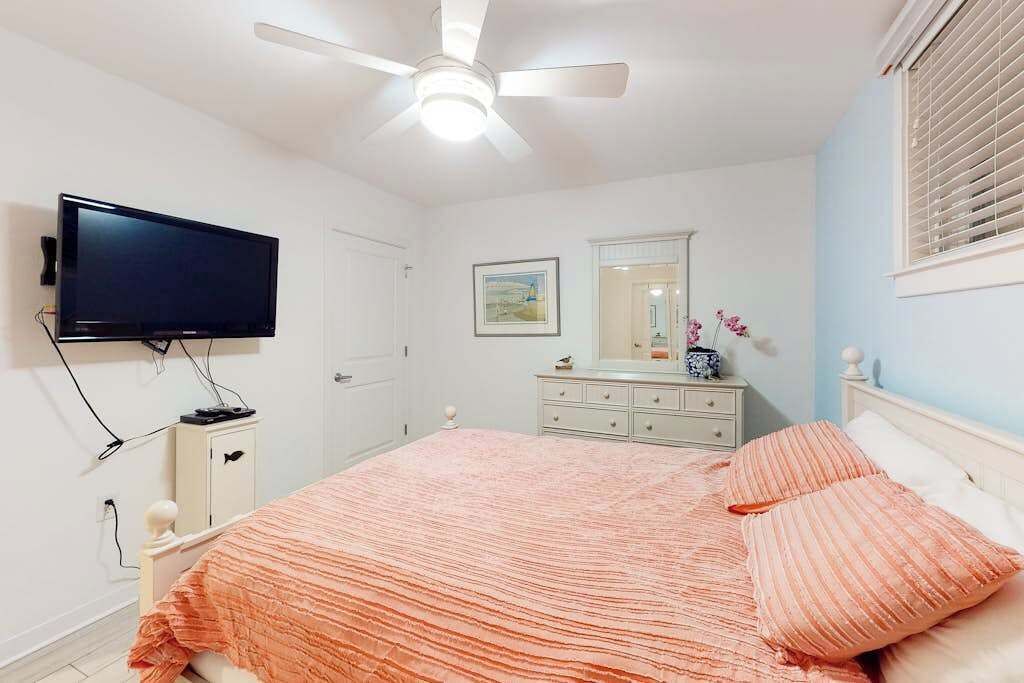 Ein Schlafzimmer mit einem Bett, einem Deckenventilator, einem an der Wand montierten Fernseher, einem Schminktisch und einem Fenster mit Jalousien.