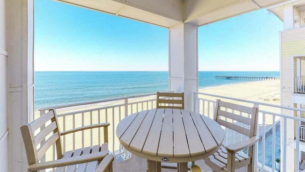 A covered balcony with a table and chairs, overlooking a beach and pier. - Oceanfront, 2-story penthouse with 3 balconies, 3 pools & 2 gyms (Virginia Beach)