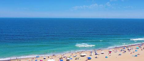 A beach with a clear sky, calm sea, and numerous umbrellas and beachgoers.