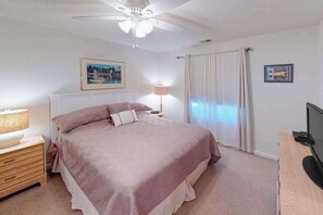 A bedroom with a bed, a nightstand, a lamp, a ceiling fan, a TV, and a window with curtains. - 1st-floor Seaside condo 1 block from beach with pool & private deck (Virginia Beach)