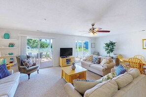 A living room with a sofa, armchair, TV, and a ceiling fan. - 1st-floor Seaside condo 1 block from beach with pool & private deck (Virginia Beach)
