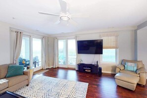 Ein geräumiges Wohnzimmer mit Deckenventilator, Flachbildfernseher, einer Couch, einem Sessel und einem großen Fenster mit Jalousien.