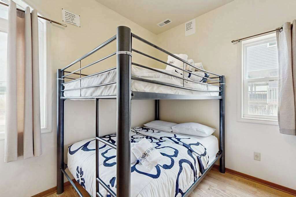 A bunk bed with a metal frame, white bedding with blue patterns, a window with curtains, and a wall-mounted sign.