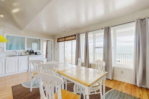 Ein Essbereich mit Holztisch und weißen Stühlen, eine Küche mit weißen Schränken sowie große Fenster mit Vorhängen.