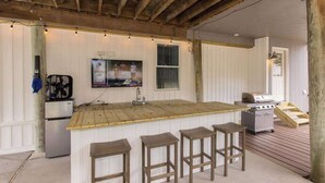 Une cuisine avec un îlot central en bois, des tabourets de bar, un réfrigérateur, un barbecue et un téléviseur fixé au mur.