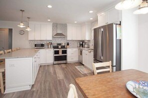 A modern kitchen with white cabinets, stainless steel appliances, and a wooden dining table. - Oceanside beach house w/game room, pool & W/D (Virginia Beach)