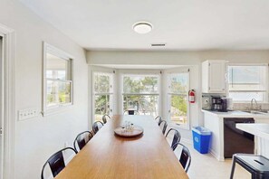 Une cuisine moderne avec des meubles blancs, une table à manger en bois et une vue sur l’extérieur grâce à de grandes fenêtres.