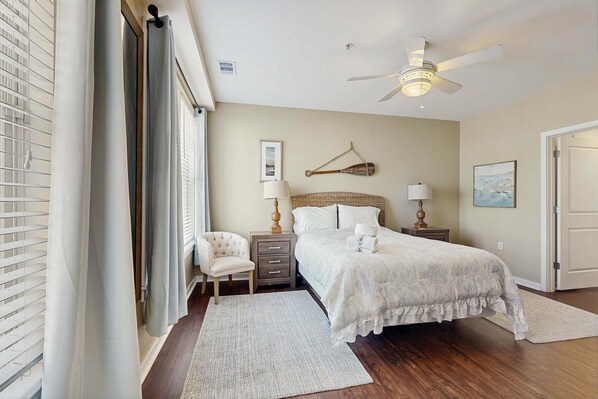 A bedroom with a bed, bedside tables, a chair, a ceiling fan, and a door. - 2nd-floor bay- & ocean-view condo with balcony, outdoor pool, grills & gym (Virginia Beach)