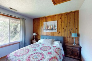 Ein Schlafzimmer mit einer holzverkleideten Wand, einem Bett mit floralem Quilt, einem Gemälde mit Segelbooten und einem Fenster mit Vorhängen.