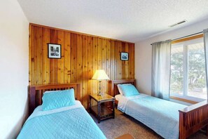 Une chambre avec deux lits, des lambris en bois sur les murs, une fenêtre avec des rideaux et une table de chevet avec une lampe.