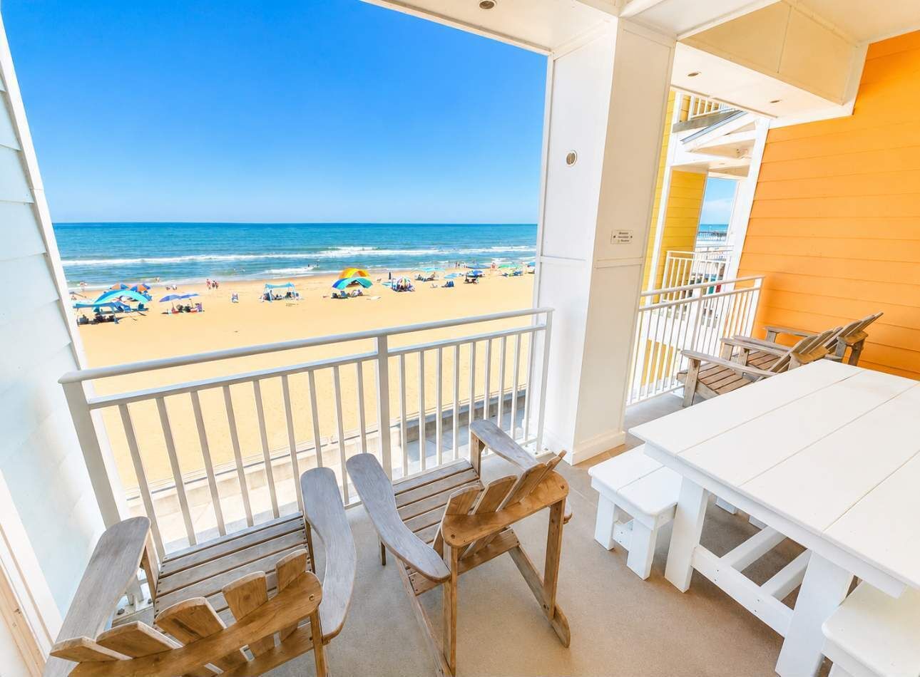 Un balcon donnant sur la plage, des chaises en bois, une table blanche et un banc blanc.