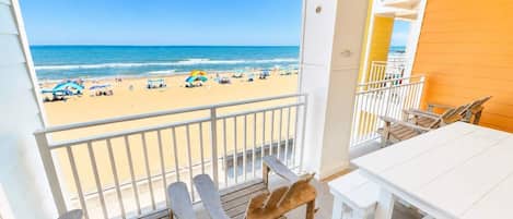 Un balcón con vista a la playa, sillas de madera, una mesa blanca y un banco blanco.