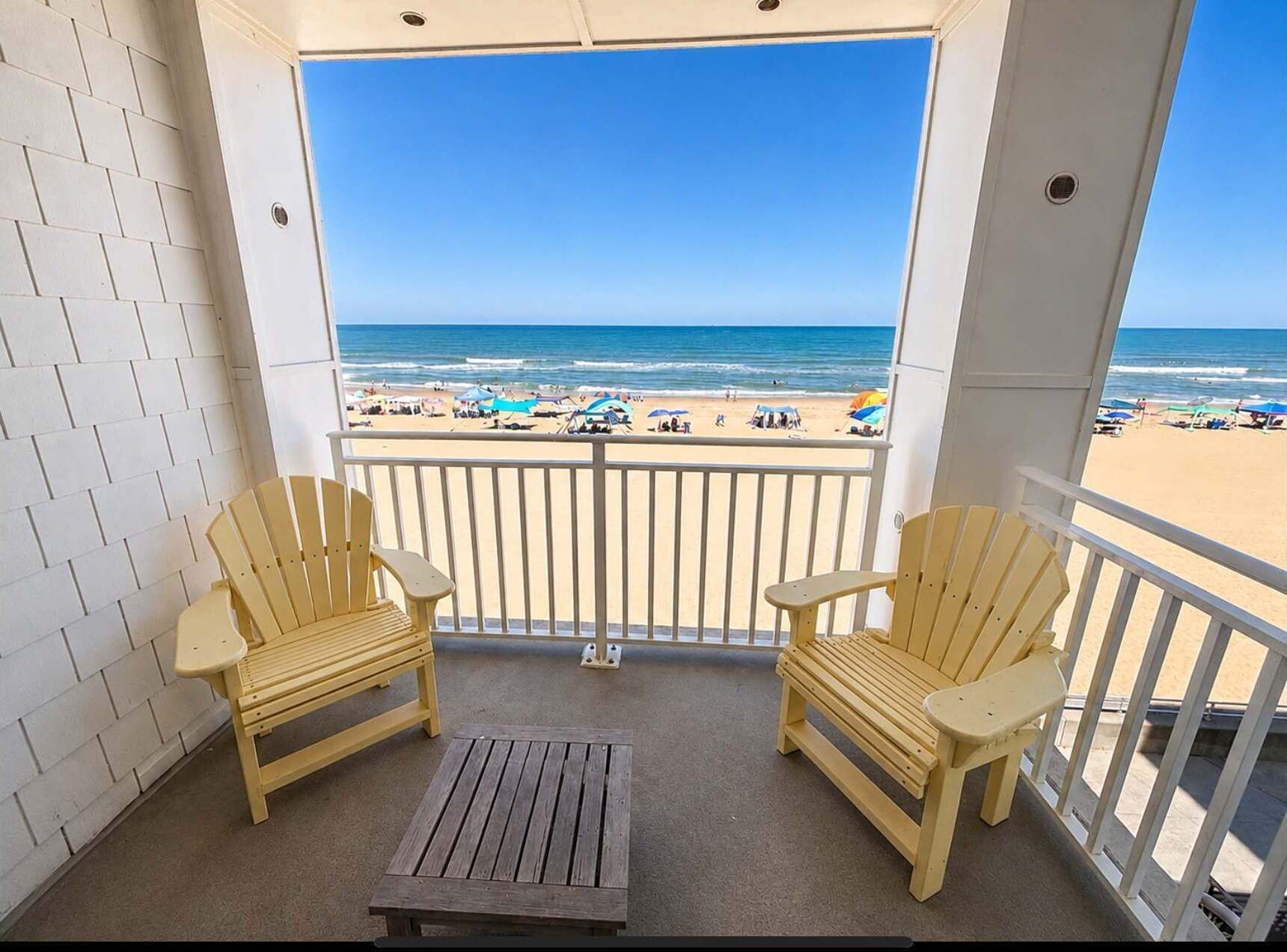 A balcony with two wooden chairs and a small table, overlooking a beach with umbrellas and people.