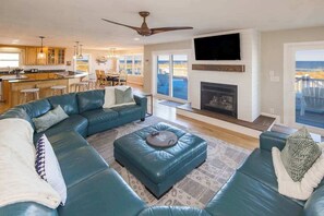 A modern living room with a sectional sofa, a fireplace, a television mounted above it, and a view of the outdoors through large windows. - Oceanfront home with private pool, crow's nest deck, game room & fireplace (Virginia Beach)