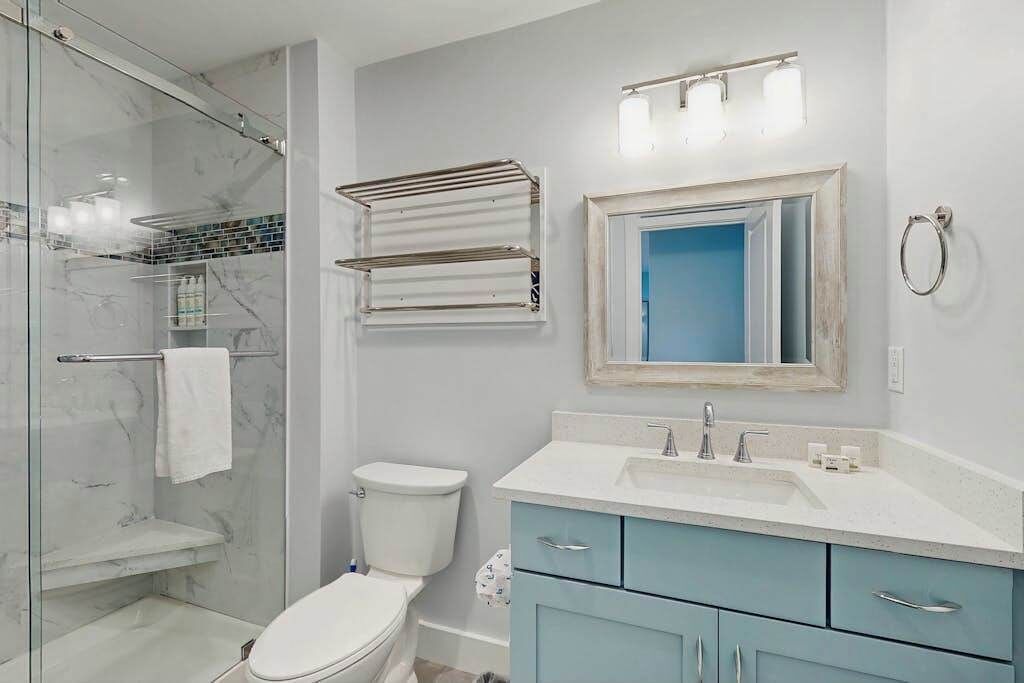 Une salle de bain avec une douche à parois de verre, des toilettes blanches, un lavabo avec un miroir au-dessus et un porte-serviettes.