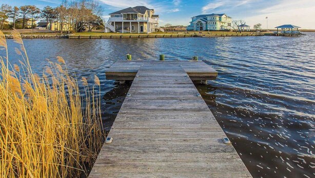 A wooden dock extending into calm water, surrounded by tall grasses and two houses in the background. - Fully-equipped canal-front home w/private outdoor pool, bay, dock, and views (Virginia Beach)