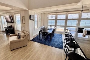 A spacious living area with a large window, a dining table with chairs, a sofa, and a flat-screen TV mounted on the wall. - Two-story, oceanfront penthouse with 2 huge balconies, 3 pools & 2 gyms (Virginia Beach)