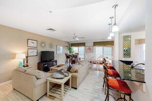 A living room with a sofa, TV, and dining area with chairs and a table. - Beachfront luxury condo with balcony, W/D, outdoor pool, gym & AC (Virginia Beach)
