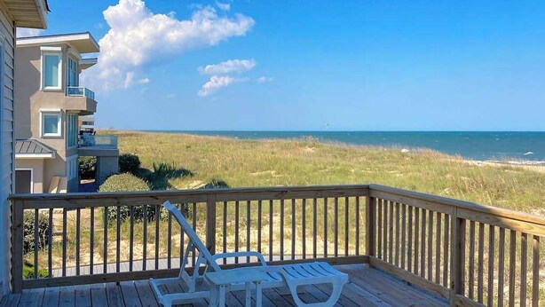 Eine Holzterrasse mit zwei weißen Liegestühlen und einem Tisch, mit Blick auf einen Strand mit Rasenfläche und Meer.