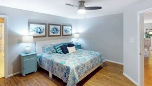 Une chambre à coucher comprenant un lit, deux tables de chevet, un ventilateur de plafond et des œuvres d’art accrochées au mur.