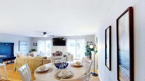 Un coin repas avec une table ronde, des chaises blanches et un bol décoratif bleu. Un téléviseur est fixé au mur, il y a un ventilateur de plafond et une fenêtre avec des rideaux.