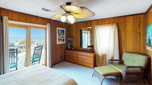 Una habitación con cama, tocador, silla, sofá de mimbre, ventilador de techo y un ventanal con vistas.