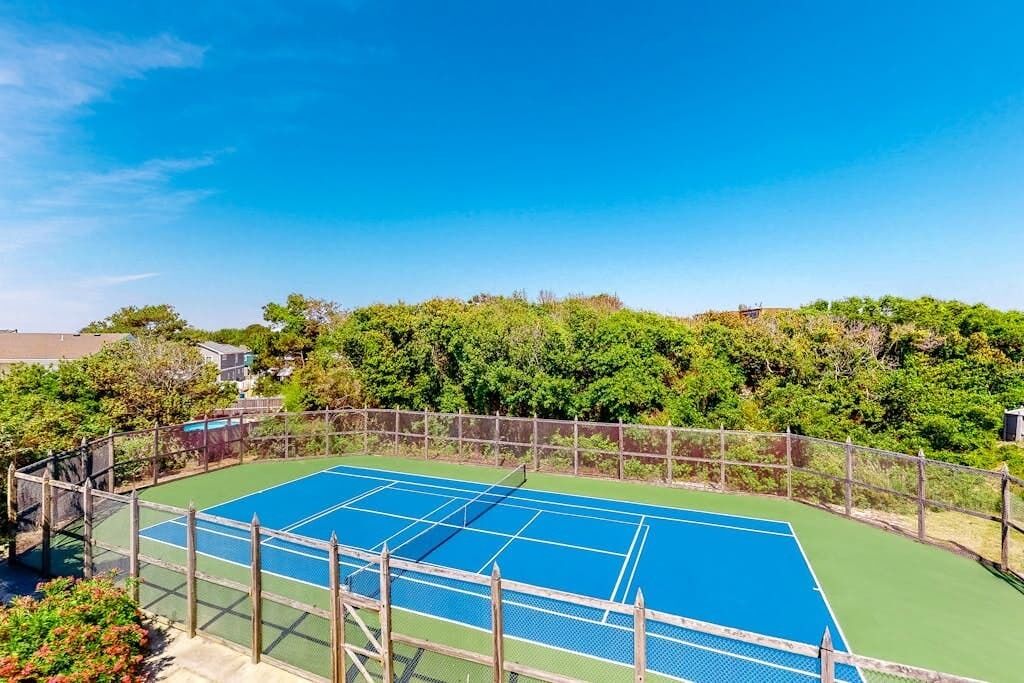 Ein Tennisplatz im Freien, umgeben von einem Holzzaun, mit dichtem Grün im Hintergrund und blauem Himmel.