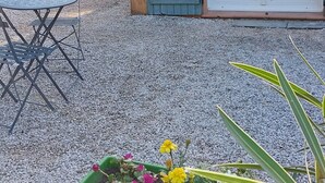 Un pequeño edificio con puerta verde y ventana, un camino de grava, una mesa con sillas y una jardinera con flores.