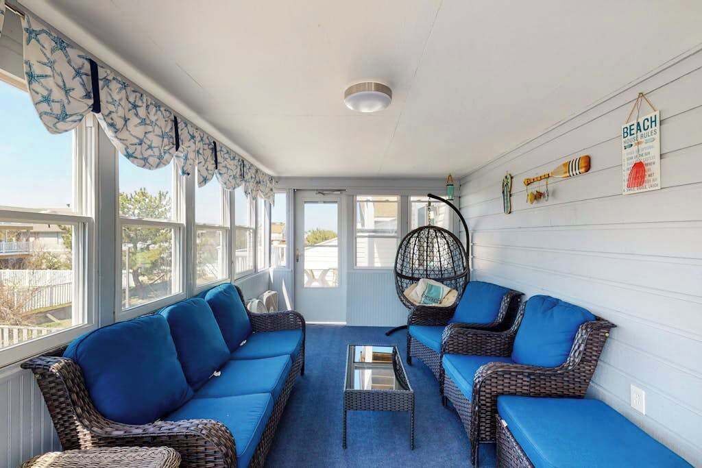 Una veranda soleggiata con mobili in vimini, un tavolino da caffè in vetro e una parete con un cartello che riporta la scritta 'BEACH HOUSE RULES'.