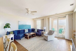 A living room with a sectional sofa, a coffee table, a side table, a lamp, a chair, and a large window with a view. - Coastal-themed beachfront condo with water views, W/D, pool, gym & AC (Virginia Beach)