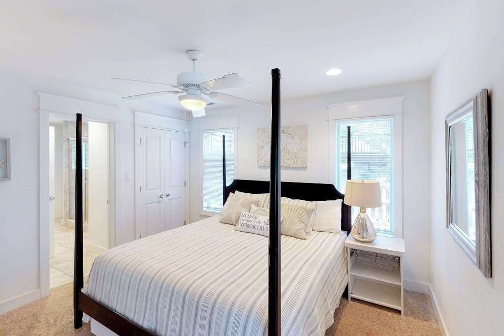 A four-poster bed with a ceiling fan, a bedside table with a lamp, and a window with blinds.