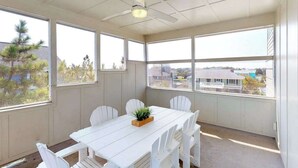 Ein Zimmer mit weißem Tisch, weißen Stühlen, einem Deckenventilator und großen Fenstern mit Blick auf Gebäude und Bäume.