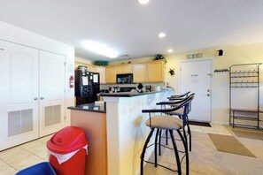 A compact kitchen with a central island, a refrigerator, microwave, and a sink. There are two bar stools and a red trash bin. - Bright Condo with On-Site Pool just a Block from the Beach (Virginia Beach)