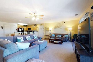A living room with a sofa, ottoman, coffee table, TV, and a ceiling fan. - Bright Condo with On-Site Pool just a Block from the Beach (Virginia Beach)