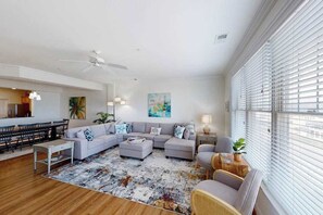 A spacious living room with a large sectional sofa, a dining area, and a kitchen in the background. - Brand-new condo steps from beach with balcony, WiFi, pool & gym (Virginia Beach)