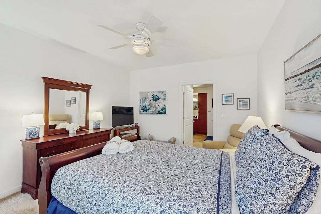 A bedroom with a bed, dresser, mirror, ceiling fan, and artwork on the walls.