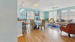 A spacious living area with a dining table, chairs, a sofa, and a TV. - Coastal corner condo with poolview balcony, resort pools & walking path to pier (Virginia Beach)
