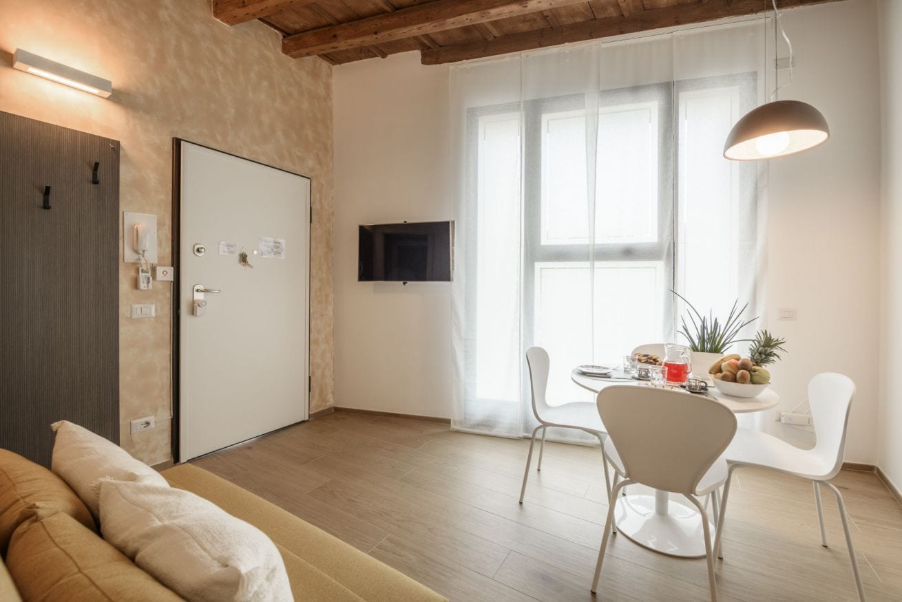 Een moderne woonkamer met een bank, een eettafel met stoelen, een aan de muur bevestigde televisie en een groot raam met dunne gordijnen.