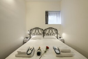 A neatly made bed with a white duvet, two pillows, and a metal headboard. There are two bedside tables with lamps, a window with a curtain, and a single red rose on the bed. - Cesariano, 11 - Downtown - Park & Movida (Milan)