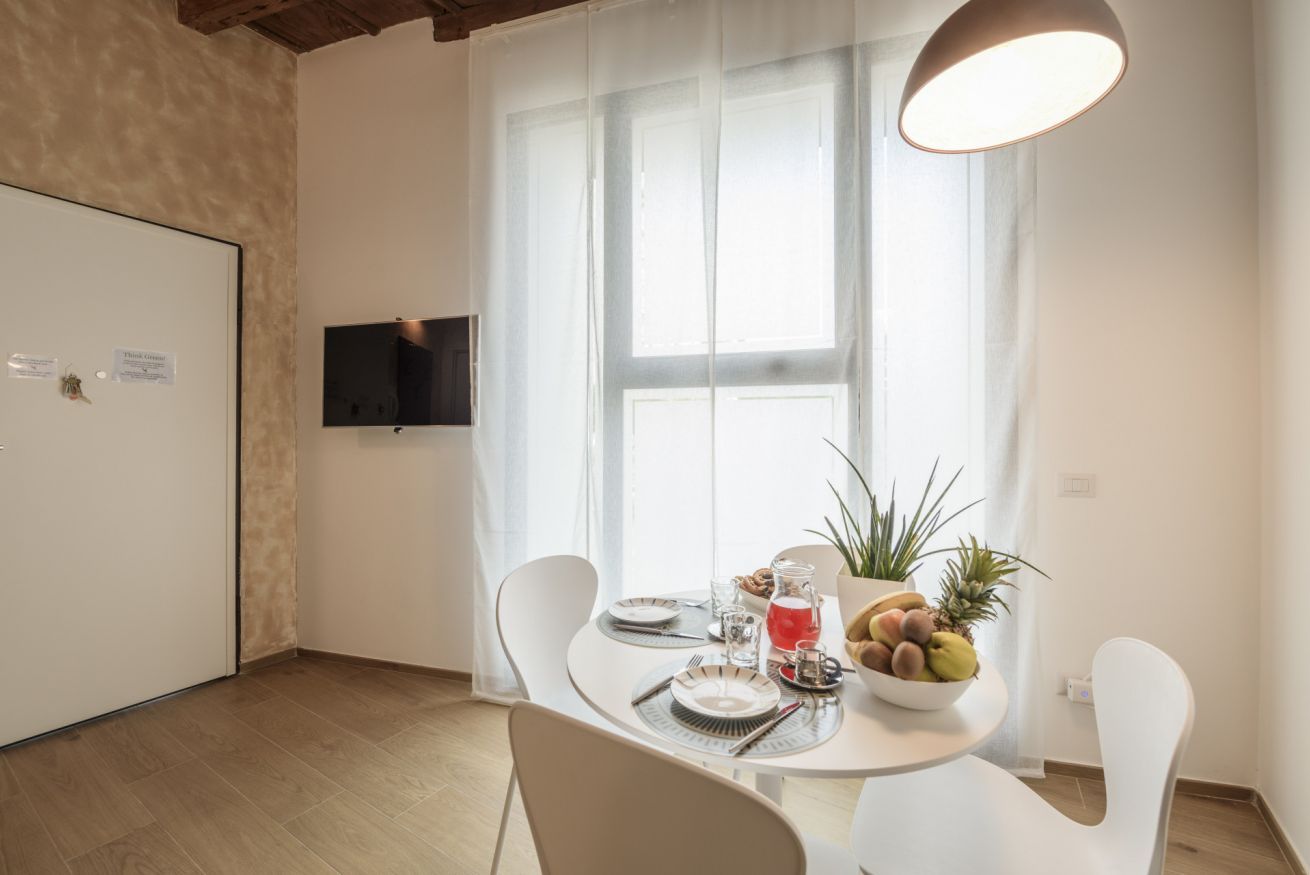 A dining area with a table set for two, a television on the wall, and a window with sheer curtains.
