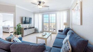 A modern living room with a sofa, a coffee table, a TV, and a balcony with outdoor furniture. - Oceanfront condo w/incredible beach views, shared outdoor pool, gym, & A/C (Virginia Beach)